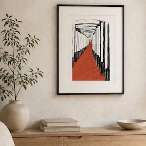 May include: A framed wall art print featuring a black and white ink-style illustration of a long corridor lined with vertical pillars, with a vibrant red path extending through the center, enclosed in a simple black frame with a white mat border.