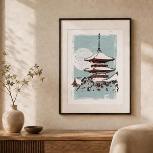 May include: A framed wall art print featuring a multi-tiered pagoda in shades of red and white against a light blue background. A large white circular moon is visible behind the structure, with dark floral elements at the base of the pagoda.