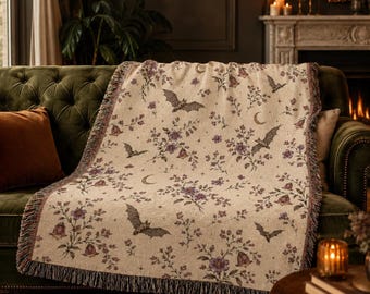 Könnte beinhalten: Eine gewebte Wohndecke mit cremefarbenem Grund und einem sich wiederholenden Muster aus fliegenden Fledermäusen, Mondsicheln, kleinen Sternen sowie violetten und grünen Blumensträußen. Die Decke hat eine strukturierte Oberfläche und Fransen.