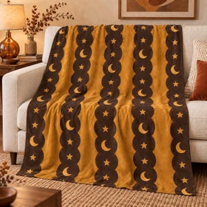 May include: A mustard yellow throw blanket featuring vertical dark brown scalloped stripes. The pattern consists of repeating crescent moon and star icons in mustard yellow printed within the dark brown bands and across the yellow sections.