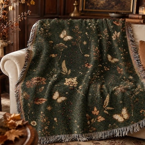 Op de afbeelding: Een donkergroene geweven plaid met een herhalend patroon van crèmekleurige vlinders, gedroogde wilde bloemen en herfstbladeren. De rechthoekige deken heeft een textuur en is afgewerkt met decoratieve franjes langs de randen.