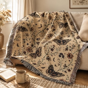 Könnte beinhalten: Eine gewebte Wohndecke in hellem Beige mit einem sich wiederholenden Muster aus dunkelbraunen Motten, Blumenranken und Mondsicheln. Die Decke hat eine strukturierte Stoffoberfläche und dekorative Fransen an den Seiten des Textils.