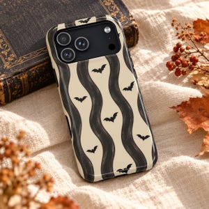 Peut inclure: Une coque de smartphone avec un fond crème orné de motifs verticaux en forme de vagues noires et de petites silhouettes de chauves-souris noires. La coque présente un bloc caméra noir avec trois ouvertures circulaires pour les objectifs.