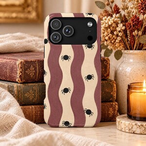 May include: A phone case featuring a repeating pattern of vertical wavy stripes in alternating shades of cream and muted mauve. Small, black, eight-legged spider icons are scattered across the surface of the case, which has a black camera cutout.