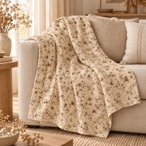 May include: A cream-coloured throw blanket featuring a repeating pattern of small brown flowers, leaves, and crescent moon shapes. The blanket is draped over the arm and seat of a light-coloured sofa, showcasing a soft, quilted-style fabric texture.