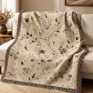 May include: A woven throw blanket in a light beige color featuring a repeating pattern of dark brown floral vines, small leaves, and crescent moon shapes. The blanket has a textured fabric finish and decorative fringed edges on the sides.