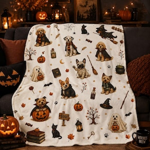 Peut inclure: Un plaid en polaire blanc orné d'un motif répétitif de chiens déguisés pour Halloween, incluant un carlin momie et un chien fantôme, avec des citrouilles, des chats noirs, des chapeaux de sorcière, des balais, des potions et des feuilles d'automne.