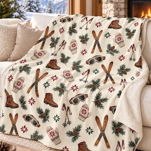 May include: A cream-colored throw blanket featuring a repeating winter pattern of brown leather hiking boots, wooden skis, ski poles, white mittens with red snowflake designs, pine branches with brown pinecones, and ski goggles.