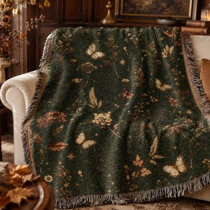 May include: A dark green woven throw blanket featuring a repeating pattern of light-coloured butterflies, various wildflowers, and scattered leaves. The blanket has a textured fabric finish and decorative fringe along the edges, draped over a sofa.
