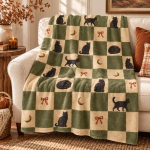 May include: A soft throw blanket featuring a checkered pattern of alternating sage green and cream squares. The design includes repeating illustrations of black cats in various poses, golden crescent moons, and small brown decorative bows.