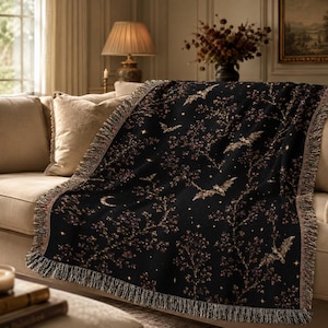 May include: A black woven throw blanket featuring a repeating pattern of flying bats, crescent moons, and small stars interspersed among dark, leafy tree branches. The blanket has a textured fringe along the edges and a soft, woven construction.