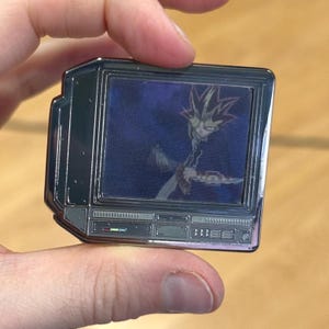 Pode incluir: Um broche de metal prateado em forma de televisão retro com uma tela azul mostrando um personagem de anime com cabelo roxo. O personagem está segurando uma espada e tem uma expressão séria.