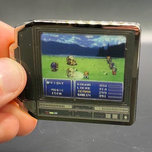May include: A black and silver pin badge in the shape of a retro television, showing a pixelated video game scene. The game features characters on a green field with text boxes. The pin is held by a hand.