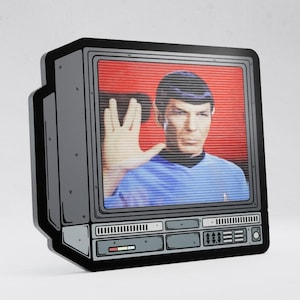 Puede incluir: Un letrero decorativo plano y rectangular con forma de televisor antiguo. La pantalla muestra el gráfico de una persona de cabello oscuro con una camisa azul haciendo un gesto con la mano sobre un fondo rojo. El marco es gris y negro.