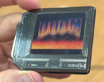 May include: A rectangular metal enamel pin featuring a dark gray frame designed to resemble a vintage computer monitor. The screen displays the word DOOM in a stylized orange and yellow font above a fiery red and orange gradient background.