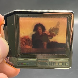 May include: A rectangular metal enamel pin shaped like a vintage computer monitor. The screen displays a printed image of a person with dark curly hair against a warm orange background. The base features detailed silver-toned control panel graphics.