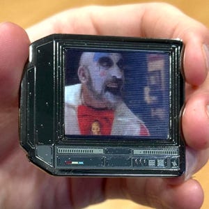 Puede incluir: Un pin de esmalte negro y plateado con forma de televisor vintage con una imagen de un hombre con pintura facial blanca y una corbata de lazo roja. El hombre lleva una camisa blanca con rayas rojas y blancas.