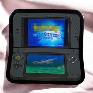 May include: A black handheld gaming console with two screens, buttons, and a directional pad. The top screen displays the text "SoulSilver Version" in blue. The bottom screen shows a scene with a body of water and a flying object.
