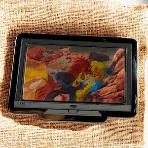 Puede incluir: Un pin negro rectangular con forma de pantalla de televisión. La pantalla muestra una escena de acción colorida con figuras de varios colores. El pin está sobre un fondo texturizado y beige.