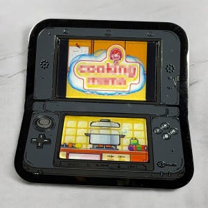 May include: A black 3DS-style enamel pin featuring a game screen with the words "cooking mama" in a cloud-shaped banner. The bottom screen shows a kitchen scene with a pot on a stove. The pin has a glossy finish.