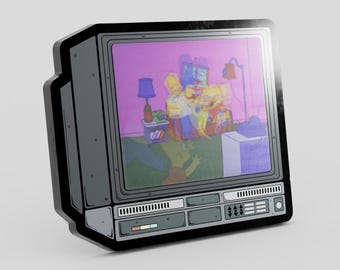 May include: A gray enamel pin shaped like a vintage television set with a screen displaying a colorful cartoon scene featuring yellow characters in a living room. The pin has a black border and detailed control panels at the bottom of the frame.
