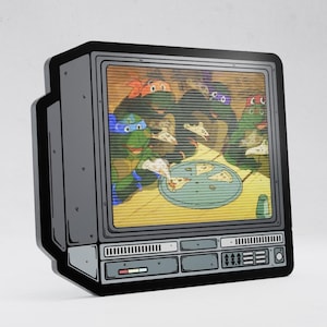 May include: A decorative wall sign shaped like a vintage television set with a black frame. The screen displays a colorful cartoon illustration of four green turtles eating pizza slices from a round tray, featuring a classic retro scanline effect.