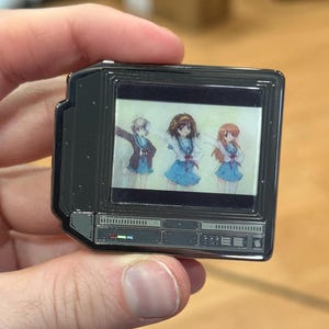 Könnte beinhalten: Eine schwarze Metallstift in Form eines Vintage-Fernsehers mit einem Bildschirm, der drei Anime-Charaktere in Schuluniform zeigt. Der Bildschirm ist von einem schwarzen Rahmen mit silbernem Rand umgeben. Die Unterseite des Stifts hat eine Reihe von Knöpfen und Knöpfen.