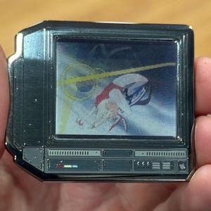 Puede incluir: Un pin de metal plateado con forma de televisor vintage con una pantalla que muestra a una mujer con un atuendo rojo y blanco cayendo por el aire. La pantalla está rodeada por un borde negro y el televisor tiene un panel de control plateado con botones y perillas.