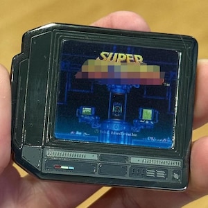 May include: A black metal enamel pin shaped like a computer monitor displaying a pixelated video game scene with the text SUPER at the top. The screen shows a central blue laboratory console with smaller monitors and the text 1994 Nintendo at the bottom.