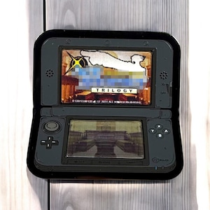 Puede incluir: Una pegatina negra con forma de consola Nintendo 3DS que muestra el juego "Trilogy" en la pantalla superior y una escena del juego en la inferior. La pegatina tiene un acabado brillante. El fondo es de madera clara.