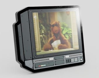 May include: A decorative enamel pin shaped like a vintage television set with a dark gray frame and control panel. The screen displays a graphic of a brown, furry creature with a long snout sitting indoors, holding a white paper object in its hands.