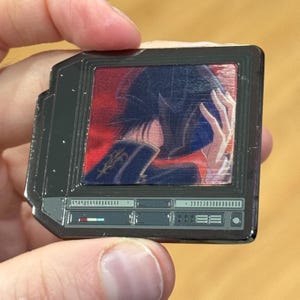 May include: A silver metal pin shaped like a vintage television with a colourful image of a person with their head in their hands. The television has a control panel with buttons and a volume knob.