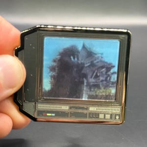 Pode incluir: Um alfinete retangular em forma de televisão vintage. A tela mostra uma imagem desfocada de um edifício e árvores contra um céu azul. O alfinete tem uma moldura dourada e detalhes em preto.