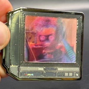 以下が含まれることがあります： ヴィンテージテレビの形をしたシルバーカラーのピン。画面には、ぼやけた、カラフルな人物の画像が表示されています。ピンには、ボタンやスピーカーなどの詳細な機能があります。ピンの端は光沢があり、反射します。