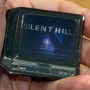 以下が含まれることがあります： 「サイレントヒル」と表示された、ビンテージテレビの形をした銀色の金属製のピン。画面は青く、光の前で立っている人のぼやけた画像が表示されています。