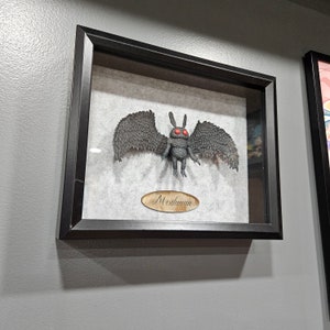 May include: Framed artwork of a Mothman figure with large, detailed wings and glowing red eyes. The dark grey figure is mounted on a light background, set within a black frame. A wooden plaque below reads "Mothman".