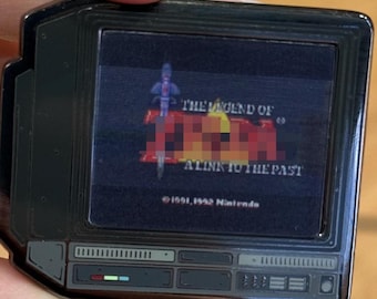 May include: A black metal enamel pin shaped like a vintage television set displaying the title screen for The Legend of Zelda: A Link to the Past. The screen shows the game logo, a sword graphic, and the text 1991, 1992 Nintendo at the bottom.