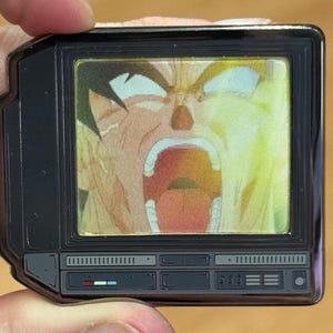 May include: A black and silver television-shaped pin featuring a cartoon image of a character with a wide open mouth and a yellow background.