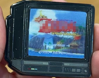 May include: A black metal enamel pin shaped like a vintage television set displaying a colorful video game scene on its screen. The screen shows a landscape with blue sky and water, and the text 2002 2003 Nintendo is visible at the bottom.