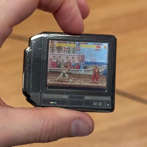 May include: A black and silver handheld video game console with a color screen displaying a video game. The screen shows a scene from a fighting game with two characters in the foreground and a background of a city street. The game's score is displayed at the top of the screen. The console has a few buttons and a directional pad.