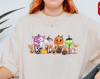 Könnte beinhalten: Elfenbeinfarbenes T-Shirt mit einem Halloween-Motiv von Bubble-Tea-Getränken. Die Grafik zeigt lila, orange und grüne Getränke mit Halloween-Dekorationen. Die Worte "Super Soft" stehen in einem roten Feld. Die Worte "more color options" stehen in einem roten Feld.
