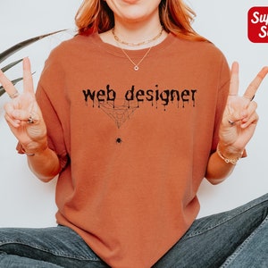 May include: A rust-colored t-shirt with the text "web designer" in black lettering with a spider web and spider graphic. The text appears to be dripping. The t-shirt is being modeled by a person wearing blue jeans.