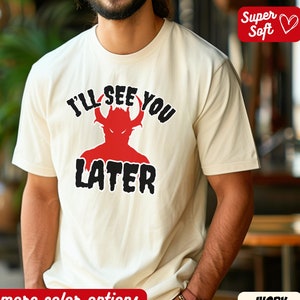 Puede incluir: Camiseta de color marfil con el texto "I'LL SEE YOU LATER" sobre una silueta de diablo rojo. La camiseta está etiquetada como "Super Soft" y "more color options".