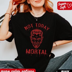 Puede incluir: Camiseta negra con el texto rojo "NOT TODAY MORTAL" y un gráfico de cara de diablo. La camiseta tiene cuello redondo y mangas cortas. Las palabras "Super Soft" están en un cuadro rojo en forma de corazón.
