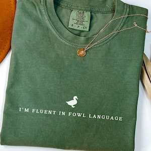 Peut inclure: Un t-shirt vert mousse avec l'inscription "I'M FLUENT IN FOWL LANGUAGE" et un dessin de canard. Un collier doré avec un pendentif repose sur le t-shirt. Un chapeau marron est visible en arrière-plan.