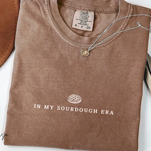 Puede incluir: Una camiseta marrón con un gráfico blanco de una hogaza de pan de masa madre y el texto "In My Sourdough Era".