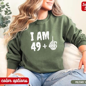 Op de afbeelding: Een groen sweatshirt met de tekst "I AM 49+" en een witte skelet hand die de middelvinger opsteekt.