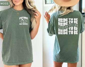 May include: Moss green t-shirt with white text on the front reading "FUTURE MRS. MEYERS" with ring graphics. The back of the shirt has the words "BRIDE TO BE" repeated in a stacked design. The shirt is a relaxed fit.