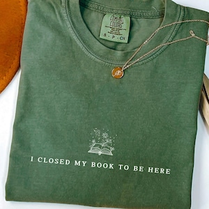 Op de afbeelding: Een groen T-shirt met de tekst "Ik heb mijn boek dichtgeslagen om hier te zijn" en een kleine illustratie van een boek met bloemen. Het shirt is gevouwen en heeft een label met de tekst "Comfort Colors" en "S-P-CH".