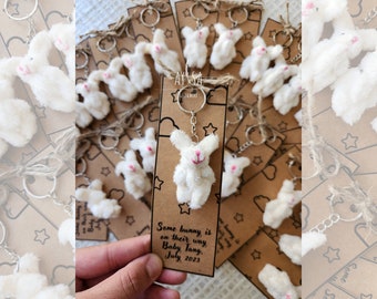 May include: A set of white bunny keychains with a brown card that reads "Some bunny is on their way Baby Tang. July 2023".